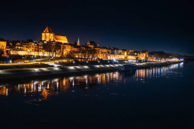 Torun 'un tarihi mimarisi gece gökyüzünün altında parlarken Vistula' nın sularında parıldayan ışıklar dans ediyor..