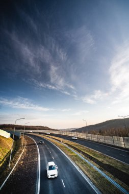 Arabalar, açık alanlar ve geniş gökyüzü ile çevrili dolambaçlı bir otoyolda dolaşarak özgürlük ve keşif hissi yaratırlar..