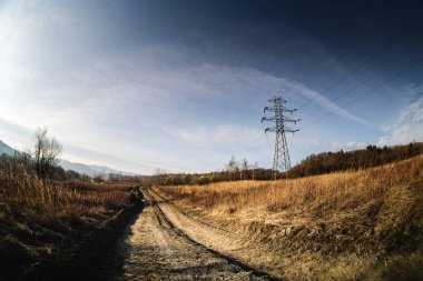 Uzun elektrik hatları, berrak mavi gökyüzünün altında altın çimenler ve yumuşak tepelerle çevrili sakin toprak bir patikanın üzerinde kule gibi..