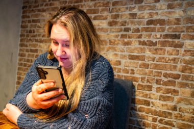 Uzun saçlı bir anne, akıllı telefonuna gömülmüş, sıcak, tuğlalı bir arka planda rahat bir ortam yaratarak köy masasında oturur..
