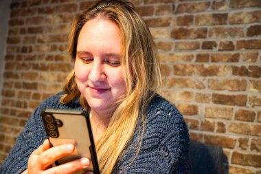 Rahat bir yetişkin kadın, sakin bir anın tadını çıkarır. Akıllı telefonuyla taşralı tuğla bir duvara yaslanır. Sıradan bir samimiyeti yansıtır..