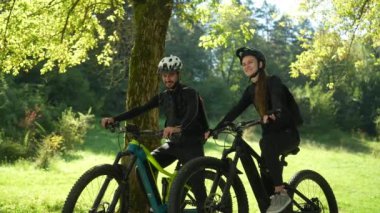 Genç Türk adam ve Bosnalı kız arkadaşı parkta keyifli bir anı paylaşıyor, bisikletlerinin üzerinde oturup ileriye bakıyor ve birlikte gülüyorlar..