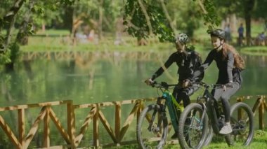 Genç adam park gölünün yanındaki ağaçların altında bisiklet sürerken kız arkadaşıyla konuşuyor..