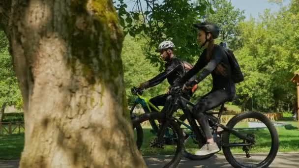公園の湖の隣の木の下で自転車に乗っている若いカップルのサイドショット — ストック動画