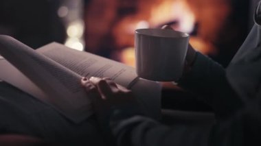 Bir insan bir fincan çay ve şöminenin yanında bir kitapla sıcak bir akşamın tadını çıkarır. Sıcak atmosfer ve huzur ortamı rahatlama ve yansıma için mükemmel bir an yaratır..