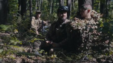 İki asker, bir keskin nişancı ve bir İHA operatörü, ağacın arkasında birbirleriyle konuşuyorlar. Üçüncü asker onlardan birkaç metre uzakta ve onları dinliyor..