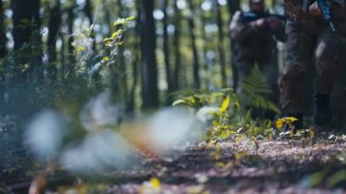 Bir askeri birlik, yoğun bir savaş sırasında yere isabet eden mermilerle, elinde tüfeklerle ormanda taktiksel olarak ilerler..