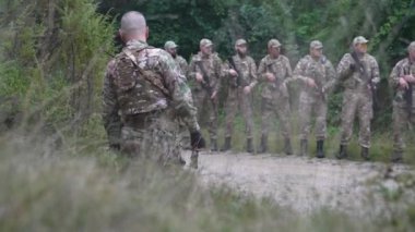 Bir askeri subay liderliğini gösteriyor. Toprak yolda askerlerini disiplinli bir sıraya diziyor. Silahlı ve hazırlıklı olan ekip, bu profesyonel açık hava eğitiminde emirleri uyguluyor..