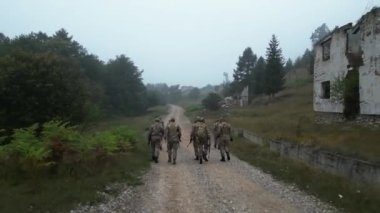 Yıkılmış evlerin yanındaki kırsal alanda, toprak bir yolda yürüyen bir askeri birliğin hava görüntüsü..