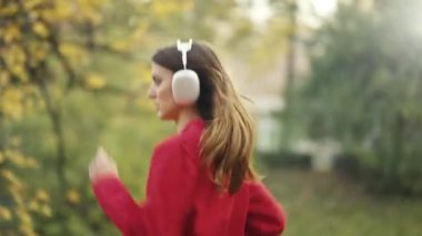 Sonbaharda elinde telefonla koşan beyaz bir kadının yan görüntüsü. Kulaklık takıyor ve ağaçların arasında koşuyor. Mevsimlik arka plana karşı aktif ve sağlıklı bir yaşam tarzı sergiliyor..