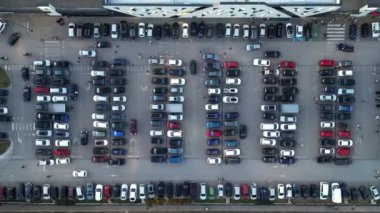 Kalabalık bir şehir manzarası. Arabalar alışveriş merkezinin dışındaki kalabalık bir otoparkta geziniyor. Tepeden tırnağa perspektif bu hareketli perakende merkezinde şehir hayatının ve tüketiciliğin enerjisini yakalıyor..