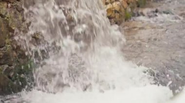 Su, eski bir taş duvarın üzerinden nehre dökülür ve bir şelale oluşturur. Kollar yavaş çekimde akar, doğanın güzelliğini gösterir. Yakın çekim, çağlayan su ve köpüğü yakalar..