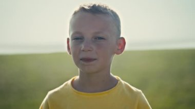 A carefree nine-year-old Bosnian boy with blond hair and blue eyes, wearing a yellow T-shirt, smiles in a close-up portrait. Set in a sunny rural meadow on a summer day in Eastern Europe.