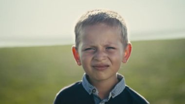 A carefree Eastern European boy with blond hair and blue eyes, squints in a close-up portrait. Set in a sunny rural meadow, the childs innocence shines on a summer day.
