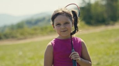 A sunlit portrait captures the joyful innocence of a five-year-old Caucasian girl. With brown hair and eyes, she smiles holding a badminton racket on her shoulder. The summer breeze is visible also.