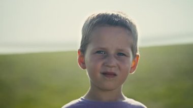 A sunny day captures the innocence of a European boy in a purple T-shirt. The close-up portrait unfolds outdoors in a rural meadow, showcasing the childs small, squinting gaze.
