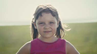 A close-up portrait captures the joy of a confident, smiling six-year-old Caucasian girl. Her brown hair waves in the summer breeze, and expressive brown eyes radiate innocence. 