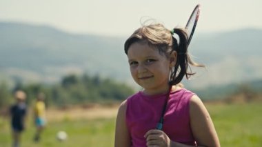 A sunlit portrait captures the joyful innocence of a five-year-old Caucasian girl. With brown hair and eyes, she smiles holding a badminton racket on her shoulder. Two boys visible behind her.