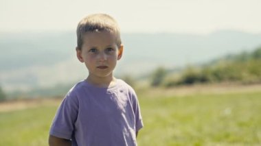 Portrait of a Caucasian child in a purple t-shirt, squinting in the bright sunlight, looking at the camera in a sunny Bosnian meadow. The small, innocent youngster in nature.
