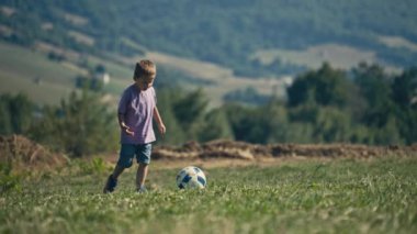 Young boy in purple T-shirt and shorts joyfully kicks a ball on green grass in nature. Active play, childhood fun, and sunny outdoor activity capture the essence of this delightful moment.