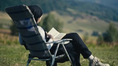 Teenage Muslim girl in nature, listing pages of a book on a picnic chair. Serene outdoor setting, hijab-clad, immersed in education and contemplation. Intellectual leisure, embracing knowledge.