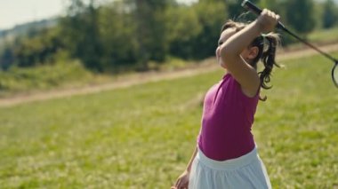 A sunlit portrait captures the joyful innocence of a five-year-old Caucasian girl. With brown hair and eyes, she smiles holding a badminton racket on her shoulder. The summer breeze is visible also.