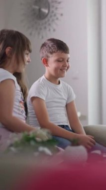 Dikey Ekran: İki utangaç çocuk kanepede oturuyor, gülümsüyor ve evde rahat bir atmosferi olan sakin bir anın tadını çıkarıyorlar. Çocukluk sevinci, kardeş bağı ve aile anları kavramı..