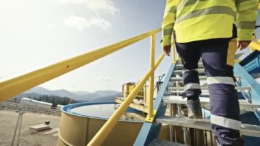 İşçi, endüstriyel bir tesiste sarı güvenlik korkuluklarıyla metal merdivenlere tırmanıyor. Yansıtıcı giysiler ve güvenlik botları giyiyor. Endüstriyel güvenlik ve açık hava çalışma ortamı kavramı.