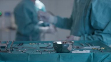 Modern bir ameliyathanede steril bir masaya yerleştirilmiş cerrahi aletlerin yakın plan çekimi. Arka planda cerrahlar bir prosedüre hazırlanıyor. Hassasiyet ve tıbbi hazırlık kavramı.