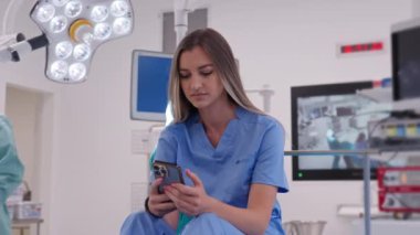 Modern bir hastane ameliyathanesinde akıllı telefon kullanan mavi önlüklü genç bir kadın doktor. Sağlık hizmetlerinde bir mola sırasında teknoloji kullanımı ve rahatlama kavramı.