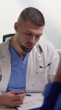 Dikey ekran: Genç doktor klinik muayenesi sırasında bir hastaya tıbbi sonuçları açıklıyor. Konuşurken doktor pano ve kalem tutuyor. Sağlık hizmetleri kavramı.