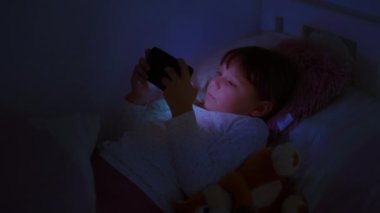 Geceleri akıllı telefon kullanan genç bir kız, mavi ışığıyla aydınlatılıyor. Ekran bağımlılığı kavramı, internet tehlikeleri ve aşırı cihaz kullanımının çocuklar üzerindeki etkisi