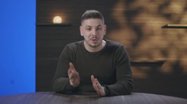 Genç adam modern bir stüdyoda bir masada oturuyor, sıcak ortam aydınlatmalı bir ortamda konuşuyor ve el kol hareketi yapıyor. Bir vlog, öğretici, içerik oluşturucu veya çevrimiçi sunum çekiliyor.