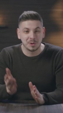 Dikey Ekran: Genç adam modern bir stüdyoda bir masada oturuyor, sıcak ortam aydınlatmalı bir ortamda konuşuyor ve el kol hareketi yapıyor. Bir vlog, öğretici, içerik oluşturucu veya çevrimiçi sunum çekiliyor.