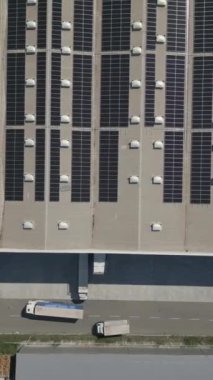 Dikey Ekran: Çatıda güneş panelleri ve aşağıda park etmiş kamyonları olan sürdürülebilir enerji ve ulaşım altyapısını gösteren bir lojistik merkezinin hava görüntüsü. Yenilenebilir enerji kavramı.