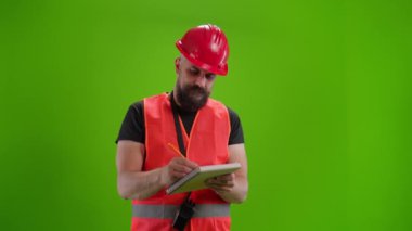Kırmızı miğfer ve güvenlik yeleği giymiş inşaat işçisi notlar alıyor, kollarını güvenle kavuşturuyor ve yeşil ekranda baş parmak işareti yapıyor. Krom anahtar, model, endüstriyel güvenlik kavramı.
