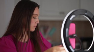 Ev ortamında video kaydeden genç bir kadın, halka ışığıyla aydınlatılmış, odaklanmış ve profesyonel görünüyor. Dijital içerik oluşturma kavramı, çevrimiçi öğretiler veya video bloglama.