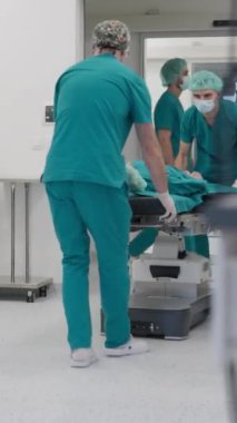 Dikey Ekran: Cerrahi önlüklü tıp uzmanları bir hastayı modern bir ameliyathane ortamında dikkatlice bir hastane yatağına taşıyorlar. Sağlık hizmetleri ekip çalışması, cerrahi hazırlık kavramı.