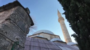 Bosna-Hersek 'in Mostar kentindeki tarihi bir camiinin, temiz bir gökyüzü altında selvi ağaçlarıyla çevrili taş bir duvar ve minareyle alçak açılı manzarası. İslam mimarisi kavramı, Osmanlı mirası.