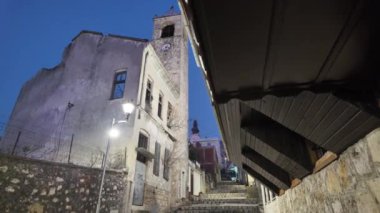 Bosna-Hersek 'in Mostar kentindeki tarihi caddede, alacakaranlıkta hasarlı bir bina ve sokak lambalarıyla aydınlatılmış bir saat kulesi yer alıyor. Kentsel bozulma, tarih ve mimari miras kavramı.