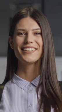Dikey Ekran: Kendine güvenen genç bir kadın gülümserken başını sallıyor, modern ofis koridorunda cam duvarlarla duruyor. Anlaşma, olumluluk ve mesleki güven kavramı.