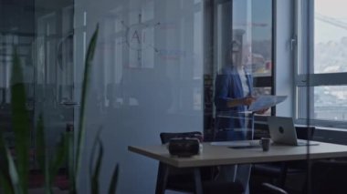 Modern bir ofisteki genç iş kadını, arka planda yapay zeka konsepti diyagramının yer aldığı cam bir tahtayla belgeleri doğal ışık altında inceliyor. Çalışma alanında teknoloji ve yenilik kavramı.