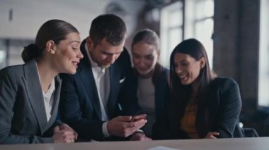 İş kıyafetleri içinde bir grup genç profesyonel modern bir ofiste akıllı bir telefona bakarken bir anlık neşe paylaşıyorlar. Kripto pazarı kavramı, borsa güncellemeleri, video izleme, video çağrısı.