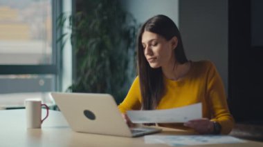 Sarı kazaklı odaklanmış genç bir kadın modern bir ofis masasındaki belgeleri incelerken dizüstü bilgisayarla çalışıyor. Profesyonel konsantrasyon ve verimlilik kavramı.