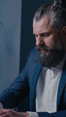 Dikey Ekran: Sakallı ve çörekli odaklanmış bir adam, evde bir mutfakta dizüstü bilgisayar üzerinde çalışıyor, takım elbise giymiş. Üretkenlik kavramı, profesyonellik ve kurumsal bir ortama odaklanma.