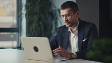 Takım elbiseli bir iş adamı profesyonel bir video görüşmesi yapıyor, modern bir ofiste dizüstü bilgisayarla yazı yazıyor ve arka planda doğal ışık ve yeşillik var. Uzaktan iletişim ve çevrimiçi çalışma kavramı.