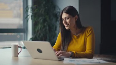 Samimi bir çalışma alanında dizüstü bilgisayarla çalışan parlak sarı bir kazak giyen ve yaratıcı bir işten zevk alan genç bir kadın. Üretkenlik, odaklanma ve modern uzaktan çalışma kavramı.