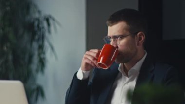 Gözlüklü profesyonel bir adam, takım elbise giyiyor, geç saatlere kadar masa başında kahve içiyor. Odaklanma, üretkenlik ve modern ofis yaşam tarzı kavramı.