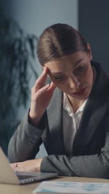 Dikey Ekran: Profesyonel iş kadını ofis ortamında bir masada oturur, dizüstü bilgisayar üzerinde çalışırken stres belirtileri gösterir ve belgeleri inceler. İşyeri baskısı kavramı.