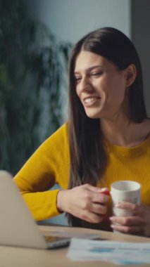 Dikey Ekran: Sarı kazaklı genç bir kadın bir video görüşmesi sırasında gülümseyip el sallıyor, bir elinde kupa tutuyordu. Uzaktan iletişim, sanal toplantılar ve modern çalışma ortamları kavramı.